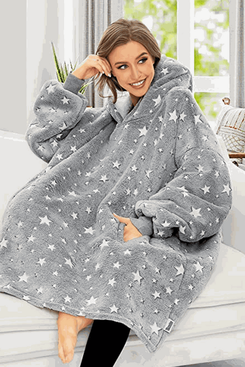 Woman wearing oversized gray Lazy Fleece Blanket Sweater with white stars, sitting indoors and smiling