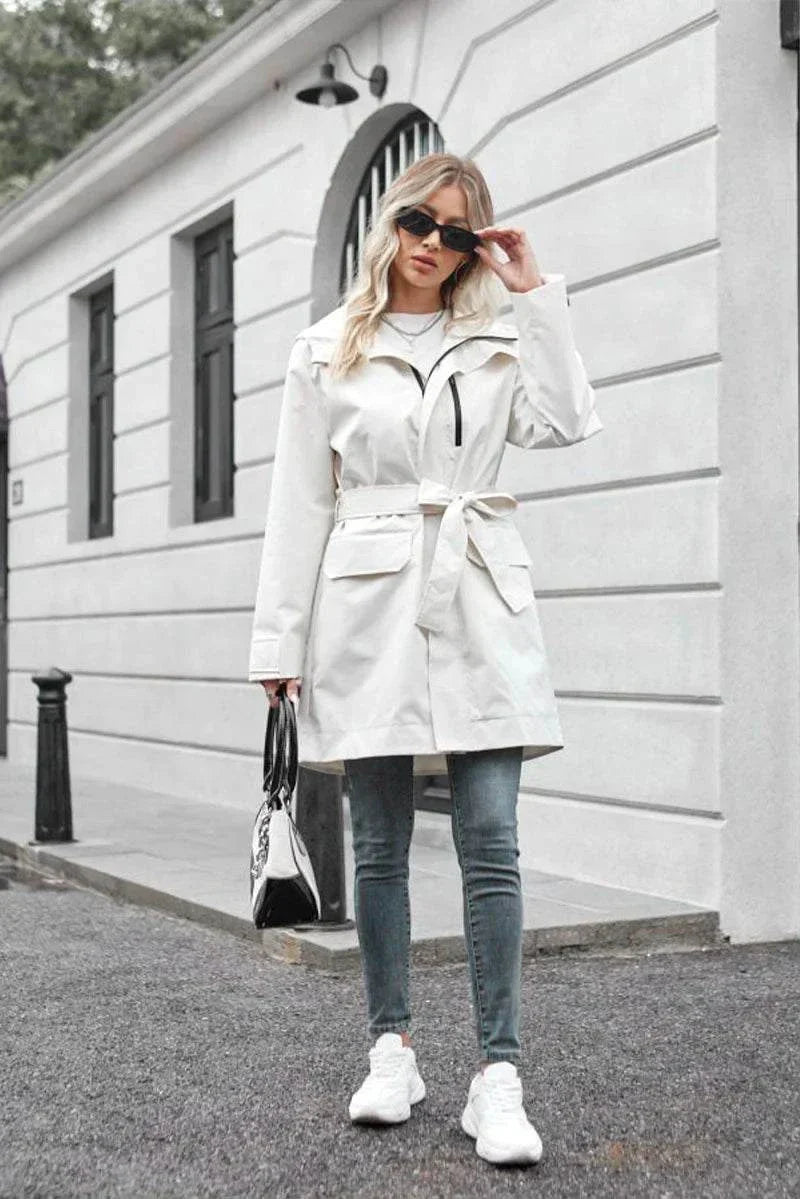 Woman wearing white water-resistant windbreaker trench jacket with belt, sunglasses, jeans, and white sneakers outdoors