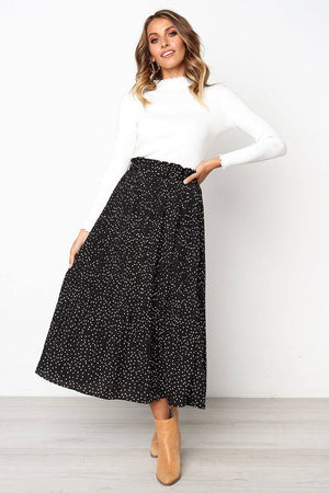 Woman wearing an effortlessly chic black polka dot pleated high-waist midi skirt with pockets paired with a white long-sleeve top and tan ankle boots