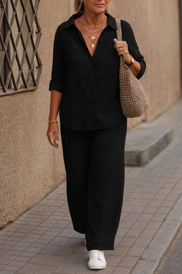 Woman wearing black relaxed shirt and trousers two-piece set with white sneakers and beige woven handbag walking outdoors