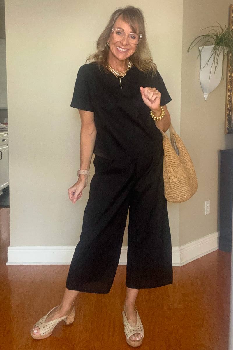 Woman wearing black loose fit co-ord set with wide short sleeves and wide-leg trousers, accessorized with necklace and straw handbag.