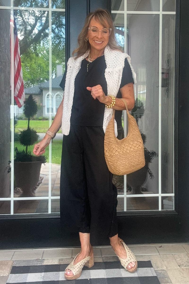 Woman wearing black loose fit co-ord set with a white knit vest, carrying a woven bag and standing in front of glass doors