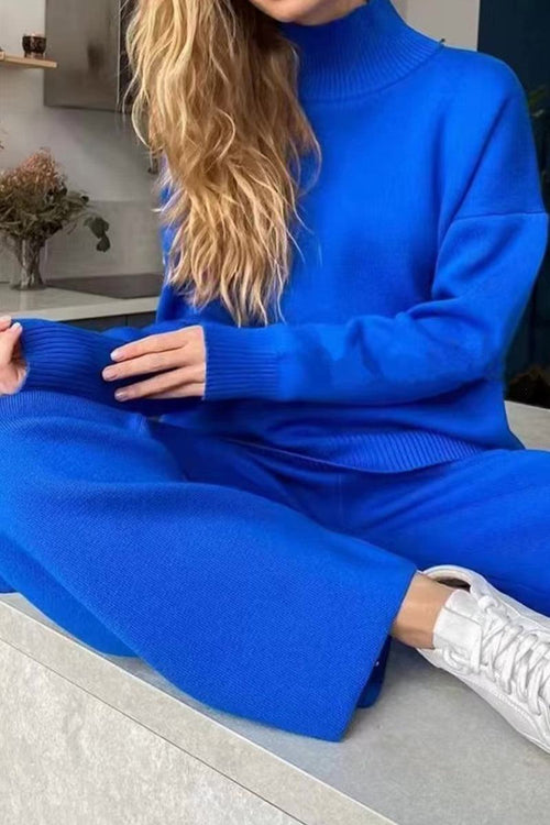 Woman wearing a bright blue knitted turtleneck two-piece set with wide-leg pants and white sneakers sitting on a ledge.
