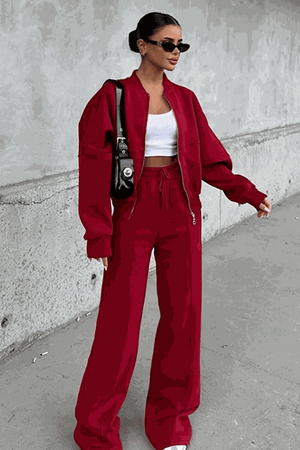 Woman wearing The Voyager Zip Set in red with wide-leg joggers and zip-front sweatshirt carrying black handbag and sunglasses outdoors against concrete wall.