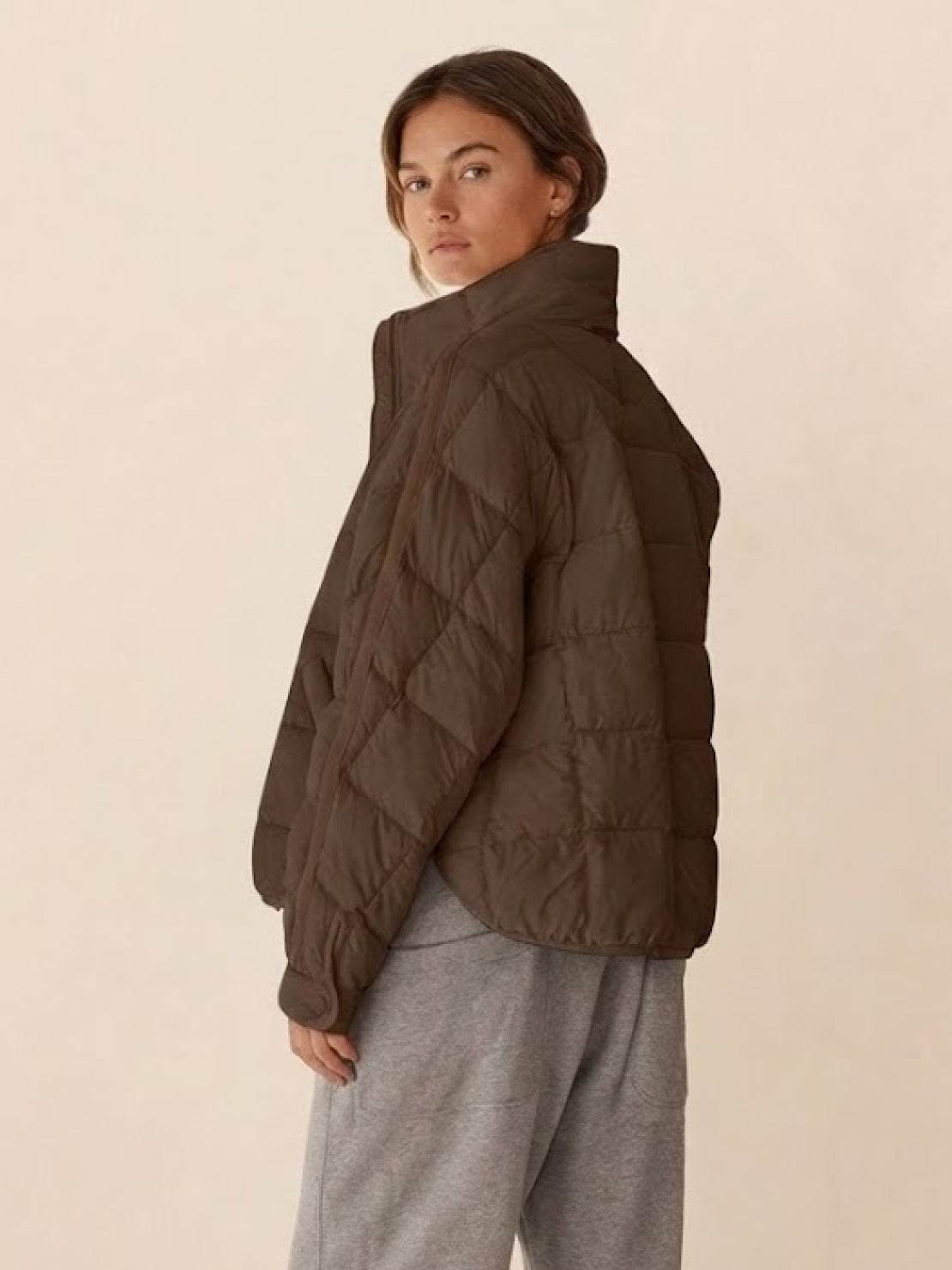 Woman wearing brown lightweight puffer travel jacket with high collar and gray pants, side view.