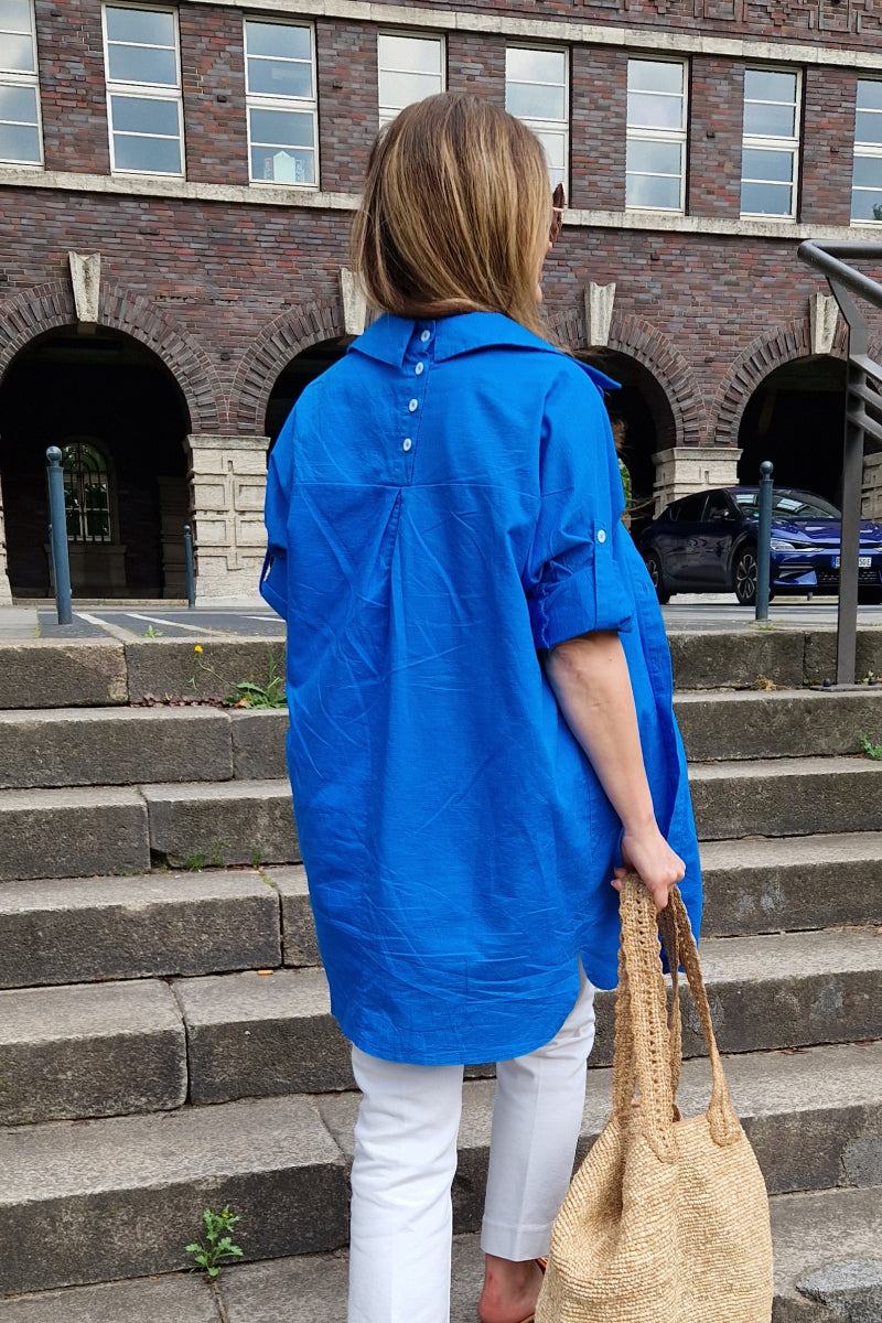 Woman wearing a bright blue oversized linen shirt with back button details walking up outdoor stone steps carrying a woven bag