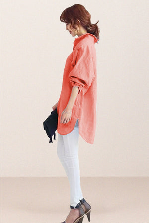 Woman wearing coral oversized linen shirt with rolled sleeves paired with white pants and heels, holding a black clutch.