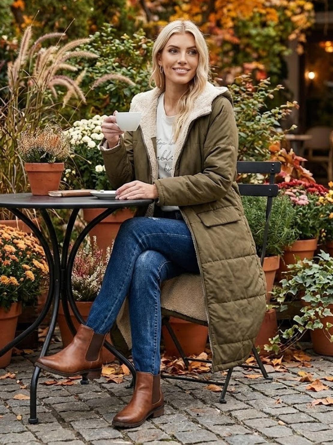 woman wearing olive sherpa-lined hooded teddy coat sitting at outdoor cafe table with fall foliage background