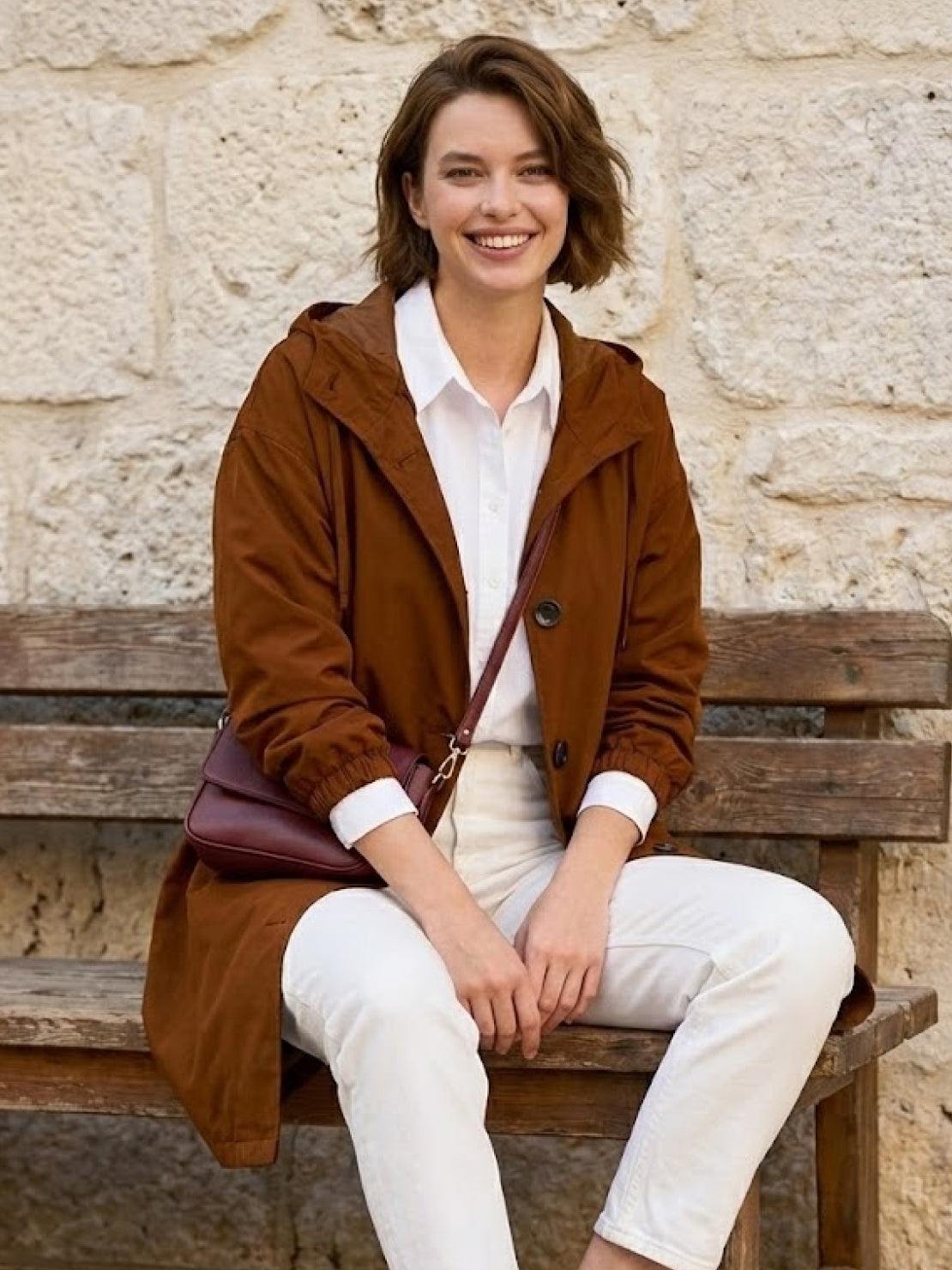 Woman wearing brown Rain Couture Windbreaker with hood, white shirt, and white pants sitting on wooden bench outdoors