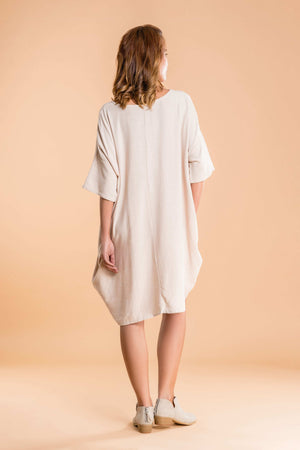 back view of nude oversized premium linen ovoid dress with short sleeves and loose fit on model against beige background