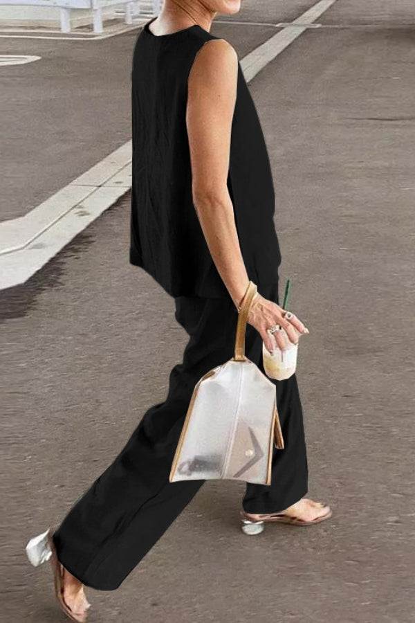 Woman wearing loose solid color black sleeveless two-piece set walking outdoors with transparent handbag and drink.