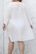 woman wearing white airy v-neck tunic with 3/4 sleeves and asymmetrical hem standing against white brick wall