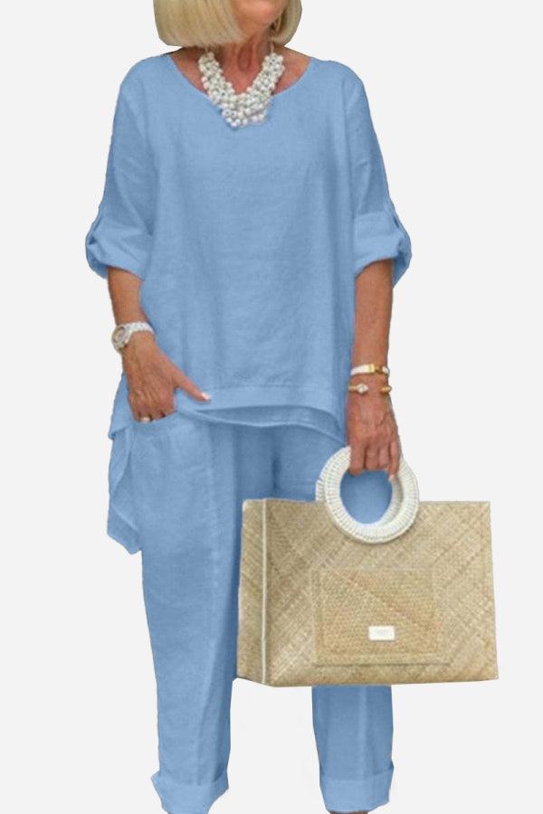 Woman wearing light blue asymmetrical two-piece trouser set with rolled sleeves and carrying beige handbag