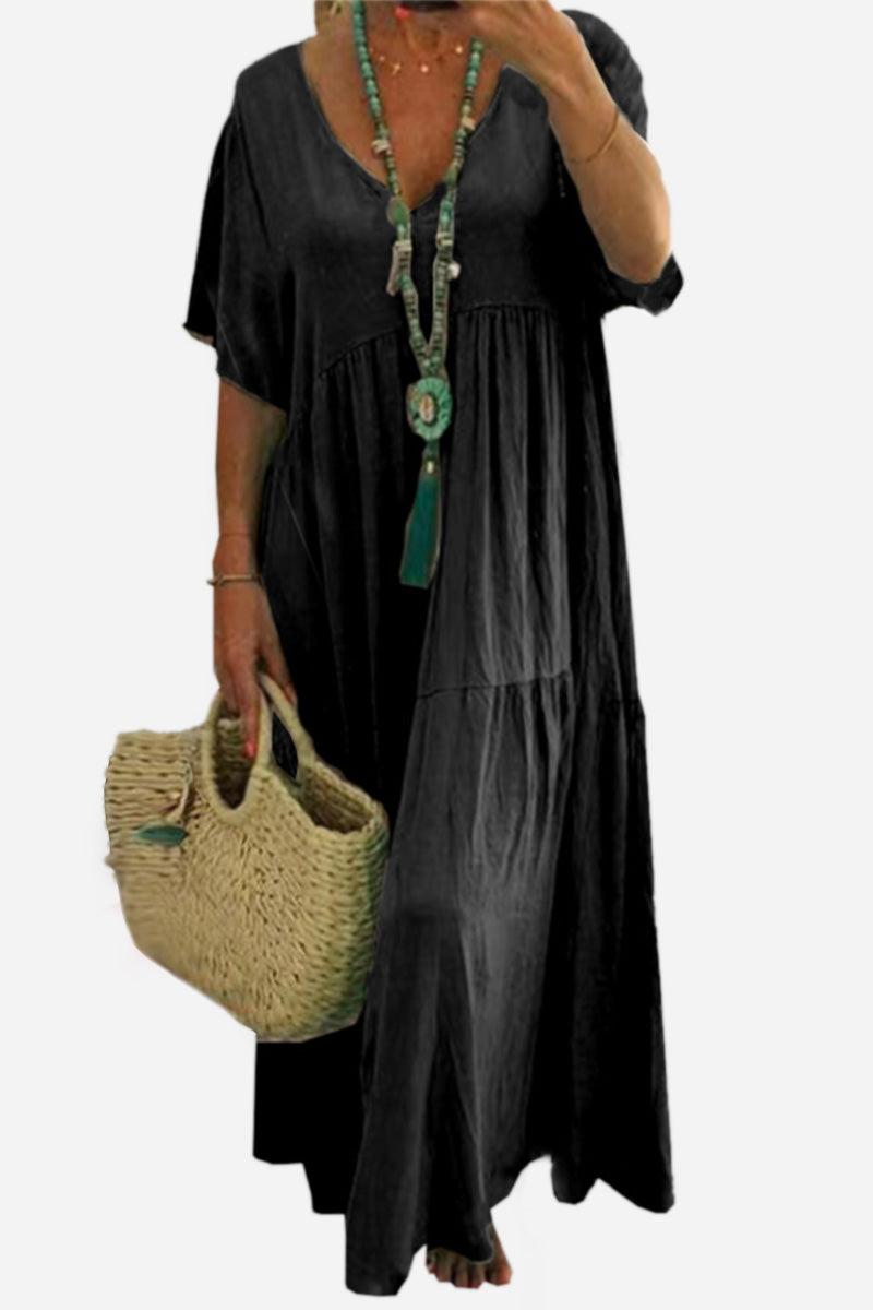 Black V-neck two-tiered maxi swing dress with loose sleeves and flowy hemline, styled with a necklace and woven handbag.