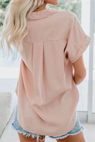 Back view of a woman wearing a light pink V-neck collared button blouse with rolled short sleeves and denim shorts.