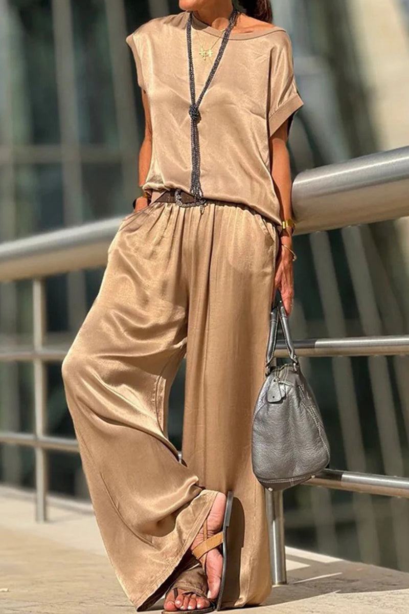Woman wearing tan Silken Escape satin two-piece set with wide-leg pants and loose top, accessorized with necklace and handbag, standing outdoors by railing