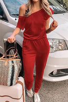 Woman wearing red Moonlit Rome Off-Shoulder jumpsuit with one shoulder exposed, standing next to a car and holding a handbag and suitcase