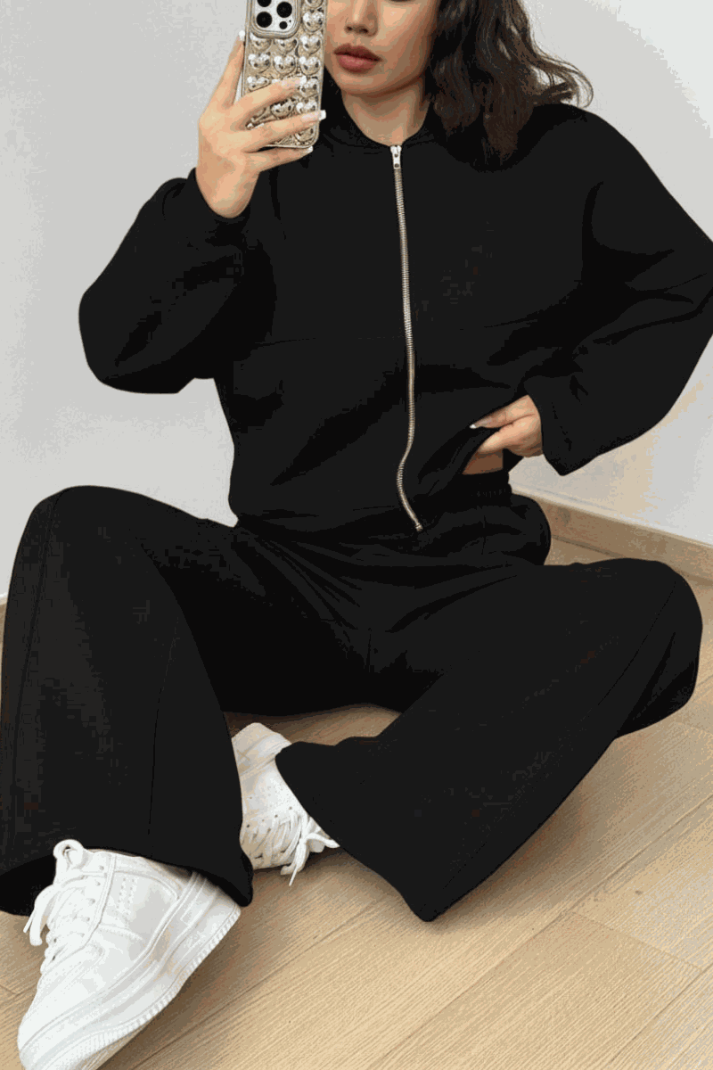 Woman wearing The Voyager Zip Set black zip-front sweatshirt and wide-leg joggers with white sneakers sitting on floor taking a mirror selfie