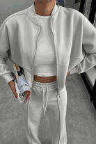 Woman wearing The Voyager Zip Set white zip-front sweatshirt and wide-leg joggers holding a magazine in a modern indoor setting