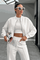 Woman wearing The Voyager Zip Set white zip-front sweatshirt and wide-leg joggers with sunglasses and holding a rolled magazine in modern indoor setting