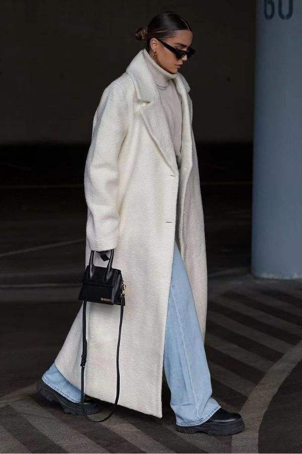 Woman wearing a long white oversized teddy coat with drop shoulders and oversized lapels, paired with light blue jeans and black boots.