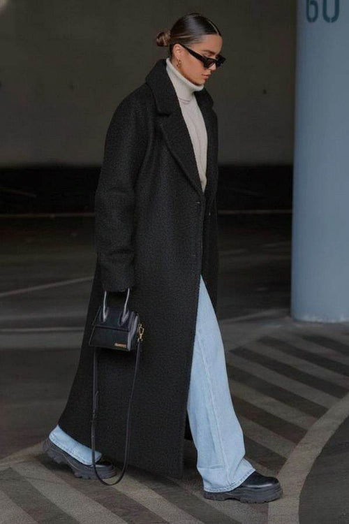 Woman wearing long black oversized teddy coat with wide lapels, light blue jeans, black boots, turtleneck sweater, sunglasses, and black handbag walking outdoors
