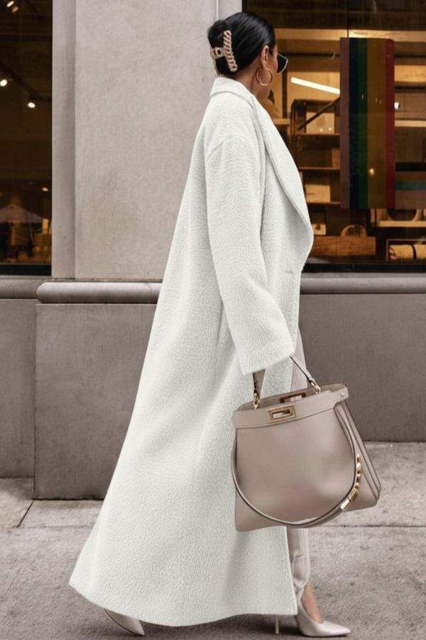 Woman wearing a white oversized teddy coat with longline silhouette and oversized lapels carrying a taupe handbag on city street