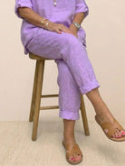Woman wearing lavender asymmetrical two-piece trouser set with rolled sleeves and tapered pants sitting on wooden stool