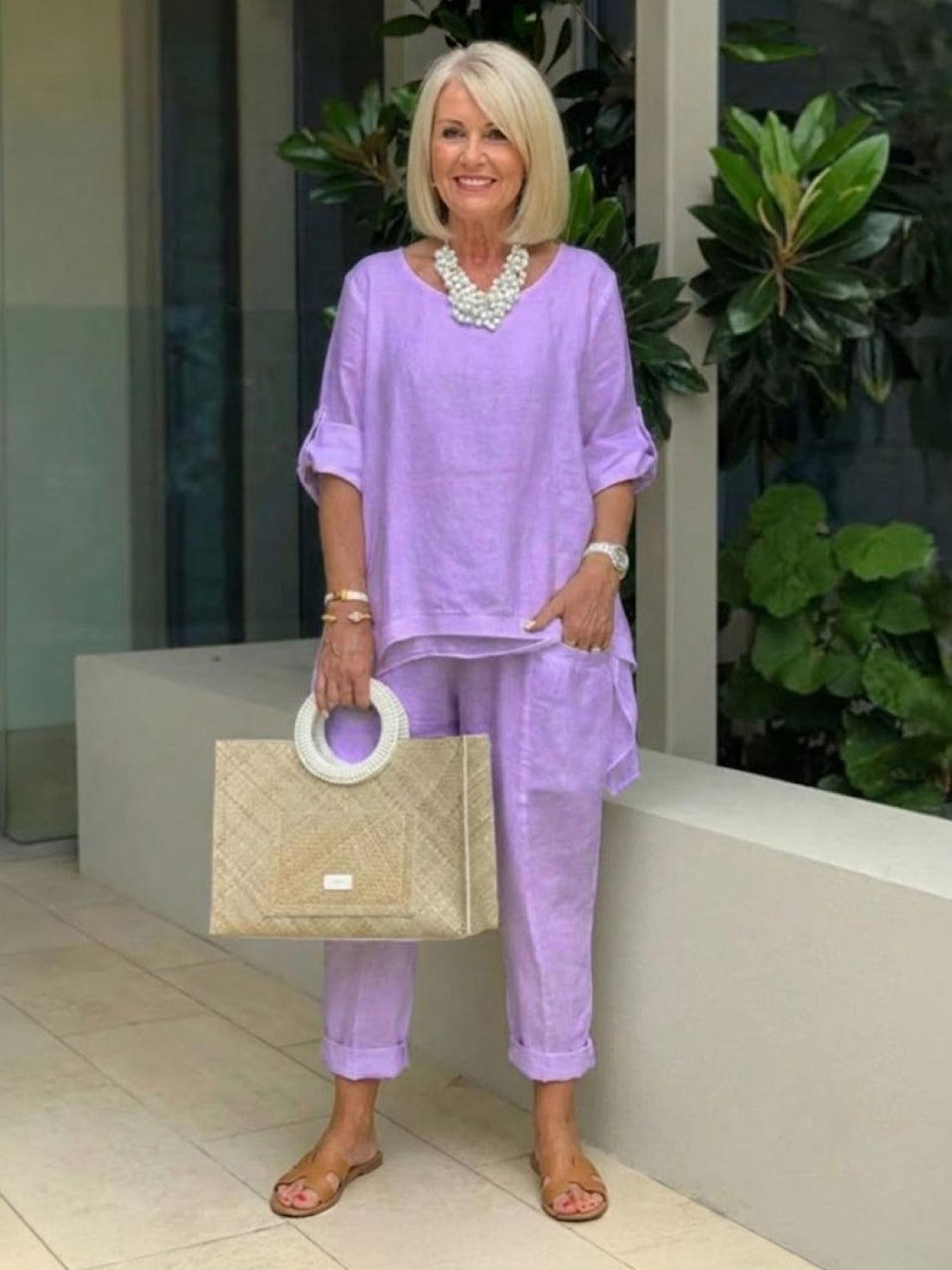 Woman wearing a lavender asymmetrical two-piece trouser set with rolled-up sleeves, paired with sandals and a beige handbag.