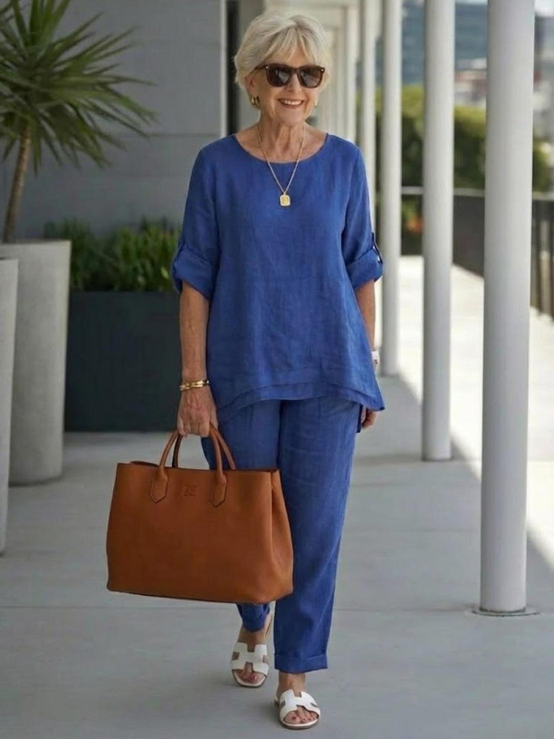 elderly woman wearing blue asymmetrical two-piece trouser set with rolled sleeves carrying brown handbag and white sandals outdoors