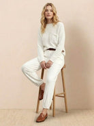 Woman wearing European minimalism relaxed knit sweater top and light drapey trousers sitting on wooden stool.