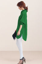 woman wearing green oversized linen shirt with rolled sleeves and white pants standing sideways holding black clutch