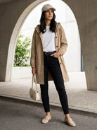 Woman wearing beige Rain Couture Windbreaker with hood, white t-shirt, black pants, beige cap, and beige loafers carrying light beige handbag.
