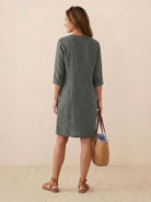 Back view of woman wearing knee-length green cotton dress with rolled ¾ sleeves and carrying straw tote bag